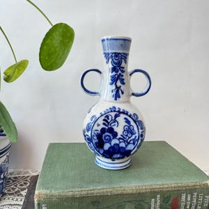 Puede incluir: Un jarrón de cerámica blanca con cuerpo redondeado y dos asas circulares. El jarrón presenta intrincados patrones florales y volutas pintados a mano en azul. Tiene un cuello estrecho y un borde acampanado, con un diseño clásico en azul y blanco.