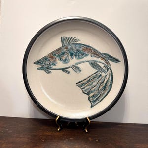 以下が含まれることがあります： 手描きの魚のデザインが施されたセラミックプレート。魚は、クリーム色の背景に青、茶色、白の色合いで描かれています。プレートは黒い縁があり、装飾的なスタンドに展示されています。