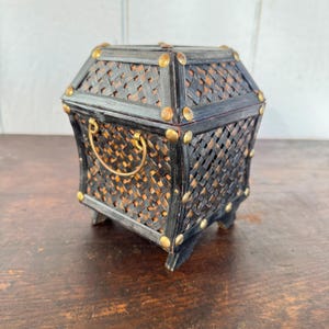 May include: A decorative black and brown woven basket with a geometric shape. The basket has a gold-colored handle and is accented with gold-colored studs. The basket is elevated on small feet and is likely used for storage or display.