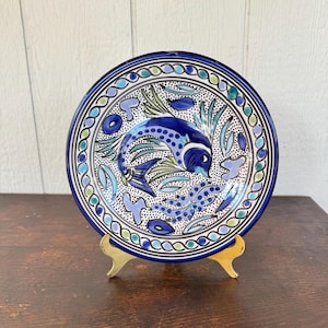 May include: A hand-painted ceramic plate featuring a blue fish design. The plate has a blue border with a repeating pattern and is displayed on a gold-colored stand. The fish is surrounded by blue and green floral elements.