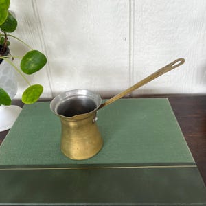 Puede incluir: Una cafetera de latón vintage con un mango largo. La olla tiene una base redondeada y un interior plateado. Se encuentra sobre un libro verde. Una pequeña planta es visible en el fondo.