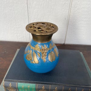 May include: A blue vase with gold-colored fruit and leaf designs. The vase has a brass-colored rim and a decorative top. The vase is sitting on top of a stack of books.
