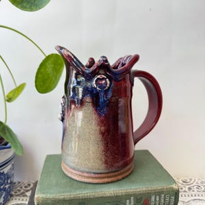 May include: A ceramic pitcher featuring a wavy, scalloped rim and a curved handle. The body displays a gradient of deep burgundy and dark blue glaze at the top, transitioning into a speckled light beige base with small white circular accents.