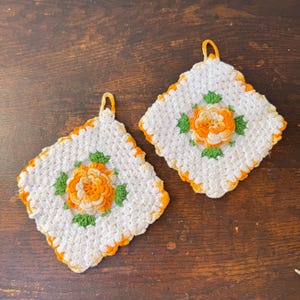 May include: Two square crochet potholders featuring a white base with a central orange flower motif and green leaves. Each potholder has an orange border and a small orange loop at the top corner for hanging, crafted with a textured yarn stitch.
