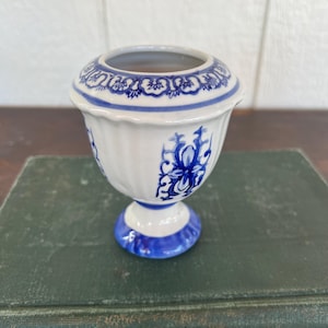 May include: A small, white ceramic vase with blue floral patterns. The vase has a rounded body, a flared rim, and a pedestal base. The blue design includes floral and scroll motifs. The vase is approximately 4 inches tall.