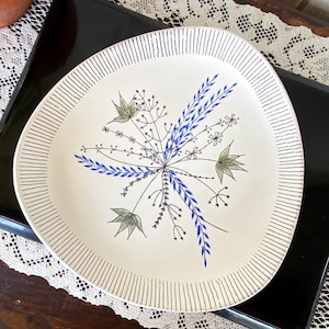 May include: Off-white ceramic plate with a unique, organic shape. The plate features a central floral design in blue, green, and black, with a striped border. A decorative piece for serving food or displaying as art.