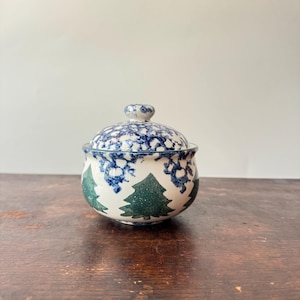 Puede incluir: Un azucarero de cerámica con tapa, que presenta una base de color crema decorada con siluetas de pinos verdes alrededor del cuerpo y un patrón de esponjado azul y blanco en la parte superior y la tapa, rematado con un pequeño pomo.