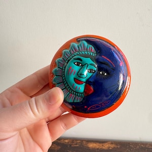 Op de afbeelding: Een keramisch decoratief stuk met zon en maan. De zon is turquoise met rode accenten en de maan is donkerblauw. De achtergrond is oranje en het stuk is rond.