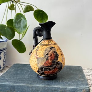 May include: A small ceramic pitcher with a black handle and neck, featuring a cream-colored body with a speckled texture. The front displays a dark brown illustration of a profile figure with detailed hair, surrounded by decorative leaf patterns.