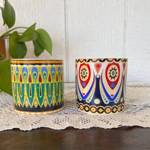 May include: Two decorative ceramic pots with colorful geometric patterns. One pot features a design with yellow, blue, and green vertical elements. The other pot has a design with red, blue, and gold shapes.