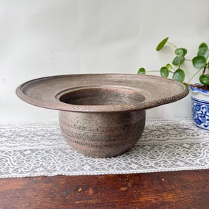 Puede incluir: Un cuenco de cobre de borde ancho con una base profunda y redondeada y un borde plano y extendido. La superficie exterior está decorada con intrincados patrones grabados repetitivos, incluyendo bandas geométricas y motivos florales.