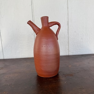 May include: A terracotta-colored ceramic pitcher with a spout and handle. The pitcher has a rounded body and a textured surface. The spout extends from the top, and the handle curves from the neck. The pitcher is displayed on a dark wooden surface.