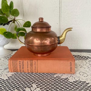 May include: A small, antique copper teapot with a brass spout and handle sits atop a vintage book. The teapot has a rounded body and a lid with a small knob. The book is orange with gold lettering.