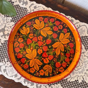 Könnte beinhalten: Runder, handbemalter Tablett mit floralem Design. Das Tablett hat einen schwarzen Hintergrund mit orangefarbenen Blättern, roten Beeren und Blumen. Der Rand ist orange mit rotem Rand. Ein dekoratives Stück zum Servieren oder Ausstellen, ca. 30 cm Durchmesser.