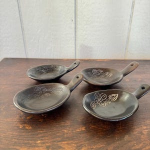 Könnte beinhalten: Vier kleine, dunkelbraune Keramik-Löffel mit Griffen, jeweils mit einem floralen Design. Die Löffel sind auf einer Holzoberfläche angeordnet und zeigen ihre einzigartige Form und dekorativen Details. Ideal zum Servieren kleiner Portionen oder als dekorative Stücke.