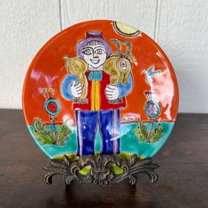 May include: A decorative ceramic plate featuring a colorful illustration of a person holding two animals. The plate has a vibrant orange background with green and blue accents. The plate is displayed on a decorative metal stand.