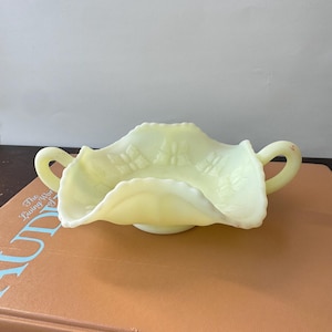 May include: A pale yellow, translucent glass bowl with two side handles and a ruffled, scalloped edge. The interior surface features a moulded relief pattern of butterflies and leaves, with a smooth, glossy finish throughout the entire piece.