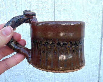May include: A handmade ceramic mug with a twisted handle. The mug is glazed in shades of brown, with darker accents near the base and handle. The mug is held by a hand, showcasing its size and shape. The mug is designed for hot beverages.