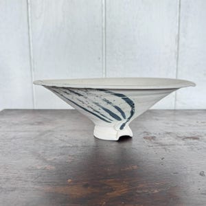 Puede incluir: Un cuenco de cerámica hecho a mano con un diseño ancho y poco profundo. El cuenco blanquecino presenta pinceladas azul oscuro y una pequeña base de pedestal blanca. El cuenco se exhibe sobre una superficie de madera.