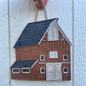 May include: A rustic, brown barn-shaped wall hanging with a dark blue roof and white accents. The ceramic piece features a detailed design of a barn with windows and doors, perfect for farmhouse decor.