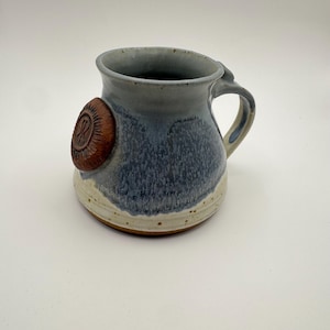 May include: A handmade ceramic mug with a blue and white glaze. The mug features a brown, circular emblem with an embossed design. The handle is a light cream color. The mug is suitable for coffee, tea, or other beverages.
