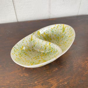 Puede incluir: Plato de cerámica ovalado con dos compartimentos. El plato es de color blanquecino con un patrón moteado de amarillo y verde. El plato está sobre una superficie de madera oscura.