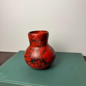 Könnte beinhalten: Eine kleine, bauchige Vase mit rundem Körper und engem Hals. Die Oberfläche ist in einem leuchtenden Rot gehalten und mit einem dichten, komplizierten Muster aus feinen, schwarzen, netzartigen Linien verziert, die das Keramikgefäß umziehen.