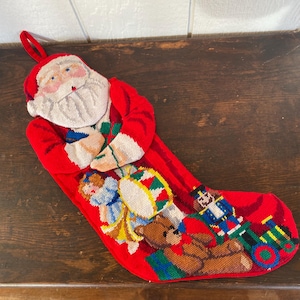Puede incluir: Un calcetín navideño rojo con la cara de Papá Noel y varios juguetes bordados. El calcetín presenta un oso de peluche, un soldado de juguete, un tambor y una muñeca. Tiene un lazo rojo para colgar.