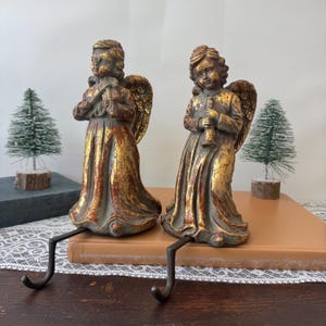 May include: Two matching stocking holders featuring standing angel figures with wings. Each angel is finished in a distressed dark grey and metallic gold paint, with one holding a small instrument. The figures are attached to dark metal hooks.