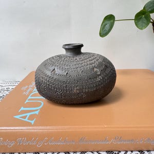 May include: A dark gray, rounded ceramic vase with a narrow neck and a wide, flattened body. The surface is decorated with intricate, repeating geometric patterns, including small circles, arches, and lines embossed across the exterior.