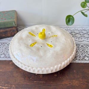 Könnte beinhalten: Ein runder, cremefarbener Keramik-Pastetendeckel mit gesprenkelter Oberfläche und gewelltem Rand. In der Mitte befindet sich ein leuchtend gelber Keramik-Apfelknauf, umgeben von fünf dampfablassenden Aussparungen in Blattform mit gelber Glasur.