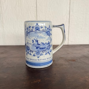 Könnte beinhalten: Ein weißer Bierkrug aus Keramik mit blauem Blumen- und Windmühlendesign. Der Krug trägt die Aufschrift "Heineken" und "Naval Station Keflavik Iceland" in blauer Schrift. Der Griff ist weiß mit blauen Akzenten.