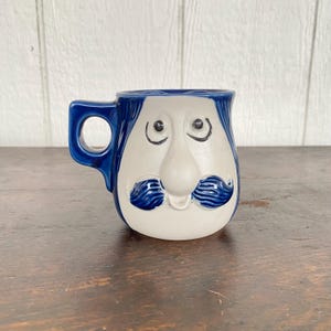 Könnte beinhalten: Eine Keramiktasse mit Gesichtsdesign. Die Tasse hat ein weißes Gesicht mit blauen Haaren, Augenbrauen und einem Schnurrbart. Der Griff und der obere Rand sind blau. Die Tasse steht auf einer Holzoberfläche.