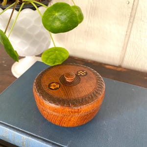 May include: A round wooden box with a lid. The box is made of a light brown wood with a darker brown decorative band around the lid. The lid has a small wooden knob and inlaid geometric designs. The box is sitting on a blue book.