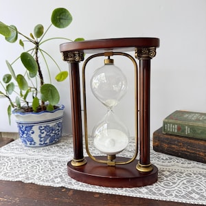 May include: An hourglass with a dark brown wooden frame featuring two columns with decorative gold-toned capitals. The central glass bulb contains white sand, held within a brass-colored metal frame that connects the oval top and bottom wooden bases.
