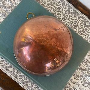 May include: A vintage copper bowl with a brass hanging loop. The bowl has a rounded shape and a polished, reflective surface. The copper has a warm, reddish-brown hue, showing signs of age and use. The bowl is resting on a teal book.