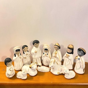 May include: A ceramic nativity set featuring figures in white robes with gold accents. The set includes Mary, Joseph, baby Jesus, shepherds, and animals. The figures have black hair and are arranged on a wooden surface.
