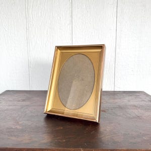 May include: A gold-colored picture frame with an oval opening. The frame is angled slightly, resting on a dark wooden surface. The background is a white wooden wall.