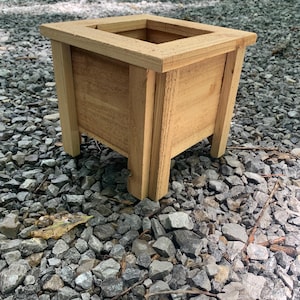 May include: A square wooden planter box with a natural finish. The planter has a rectangular opening at the top and is supported by four legs. The box is constructed from light-colored wood and sits on a bed of gray gravel.
