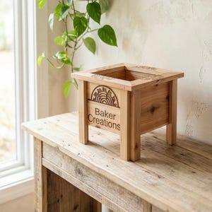 May include: A wooden planter box with the text "Baker Creations" and a sunburst design. The square planter is made of light-colored wood and sits on a rustic wooden table. A green plant is visible in the background.