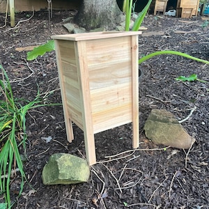 May include: A rectangular wooden planter box with a natural finish. The planter has a boxy shape with four legs and is made of light-colored wood. The planter is outdoors, surrounded by mulch and greenery.