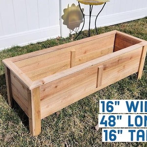 May include: A rectangular wooden planter box, 16 inches wide, 48 inches long, and 16 inches tall. The planter is made of light-colored wood and has a simple, clean design. The box is sitting on grass.