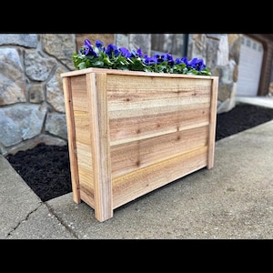 May include: A rectangular wooden planter box filled with vibrant purple flowers. The planter is constructed from light-colored wood planks and features a simple, clean design. The box is placed on a concrete surface, with a stone wall in the background.