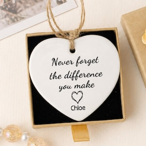 May include: A white heart-shaped ceramic ornament with a jute twine hanger, featuring the black printed text "Never forget the difference you make" and a heart icon above the name "Chloe," presented in a square cardboard gift box.