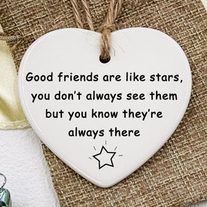 Könnte beinhalten: Ein weißes herzförmiges Keramikornament mit einer Juteschnur. Das Ornament trägt den Text "Good friends are like stars, you don't always see them but you know they're always there" und einen Stern. Es liegt auf einem Jutesack.