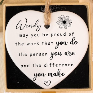 Pode incluir: Placa de cerâmica branca em forma de coração com borda preta, com o nome "Wendy" e uma mensagem inspiradora. A placa está em uma caixa de presente com uma etiqueta "Hand made". Flores amarelas e detalhes florais secos.
