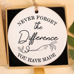 Può includere: Un ornamento a disco in ceramica bianca con la scritta "NEVER FORGET the Difference YOU HAVE MADE" in nero. L'ornamento è in una scatola nera con un nastro dorato. È visibile anche una scatola marrone con la scritta "Hand made".