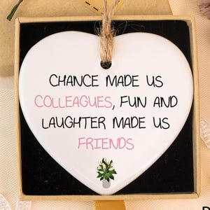Op de afbeelding: Een wit hartvormig keramisch ornament met de tekst "CHANCE MADE US COLLEAGUES, FUN AND LAUGHTER MADE US FRIENDS". Het ornament zit in een zwarte doos met een bruin deksel en een "Hand made" label. Gele bloemen liggen bovenop.