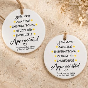 Può includere: Due ornamenti rotondi in ceramica bianca con testo nero che recita: you are AMAZING INSPIRATIONAL DEDICATED INCREDIBLE Appreciated. Sotto il testo c'è un piccolo cuore e la frase: Thank you for being a part of our team.