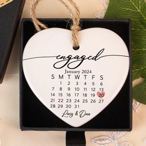 以下が含まれることがあります： 「engaged」の文字と2024年1月のカレンダーが描かれた白いハート型のオーナメント。日付の20がハイライトされています。オーナメントは「Hand made」のタグが付いた黒い箱に入っています。